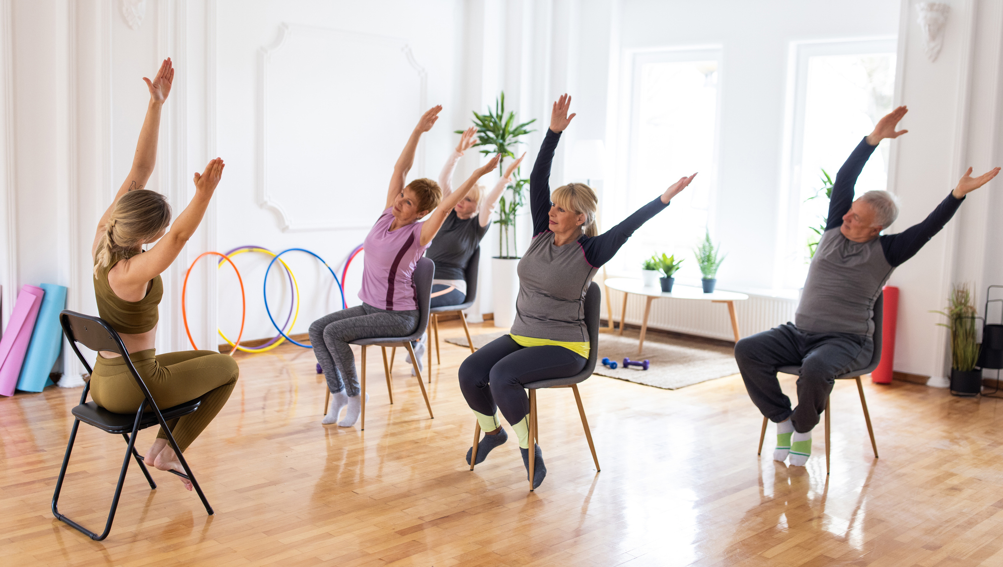 An instructor leads a seated exercise class as participants raise their arms in a bright studio with mats, hoops, and plants in the background.