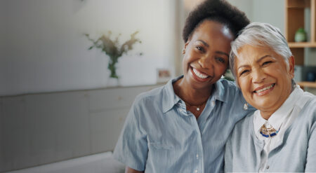 Two adults sit close together on a couch, with one person’s arm around the other. Both individuals wear light-colored shirts. Behind them is a softly lit living room with cabinets, shelves, framed photos, and plants.
